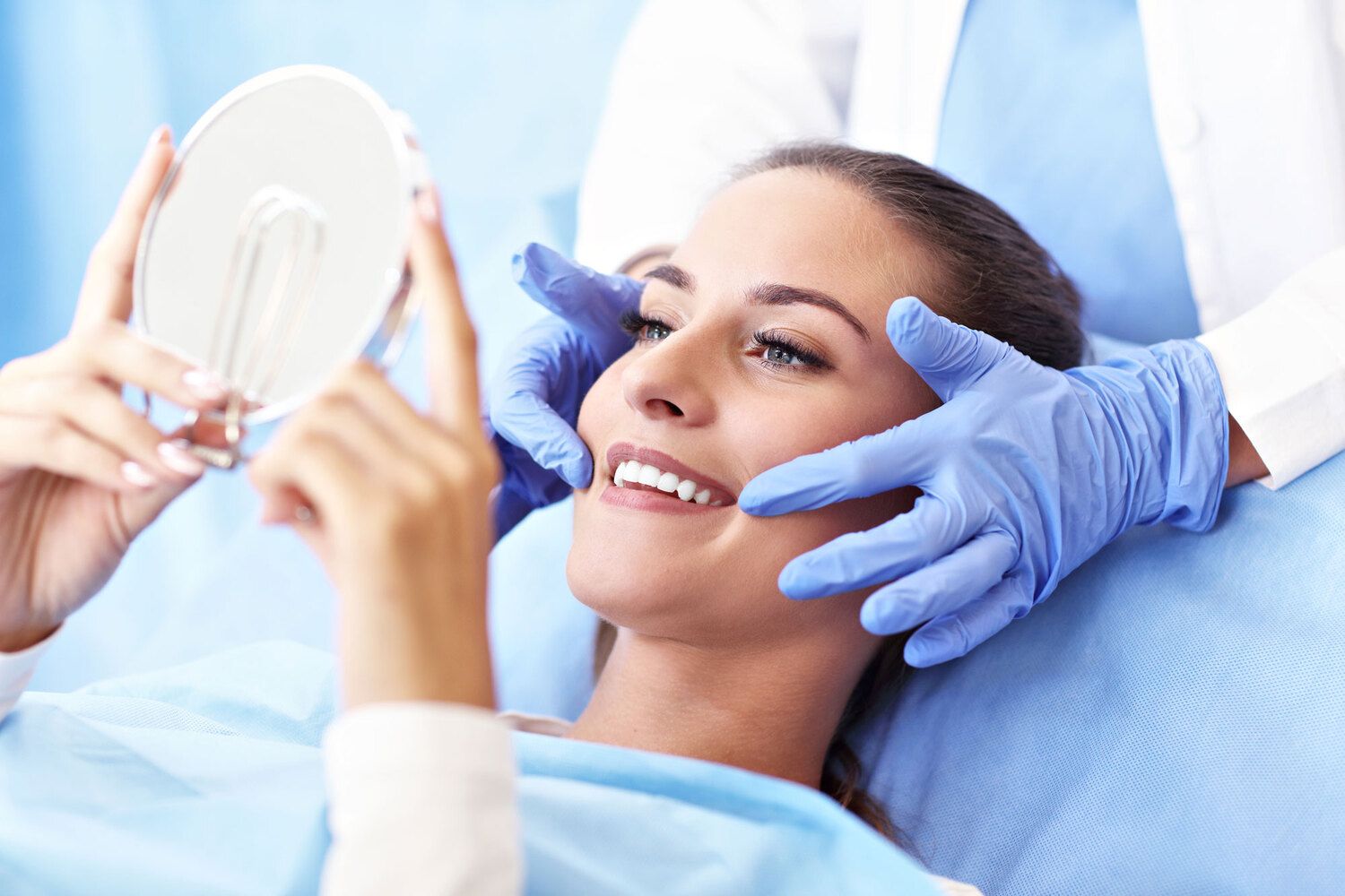 Smiling woman in a dental chair holding a mirror while a dentist in gloves examines her teeth.